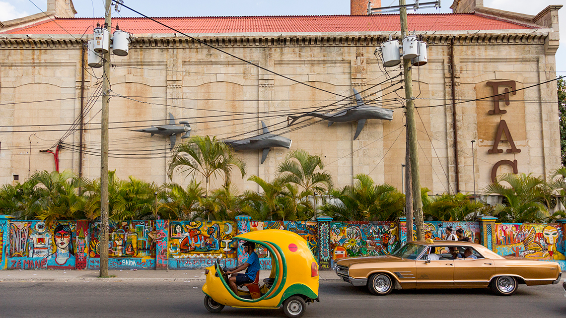 A cocotaxi and a vintage american car in the streets of Havana, in the background the FAC and its wall full of graffiti